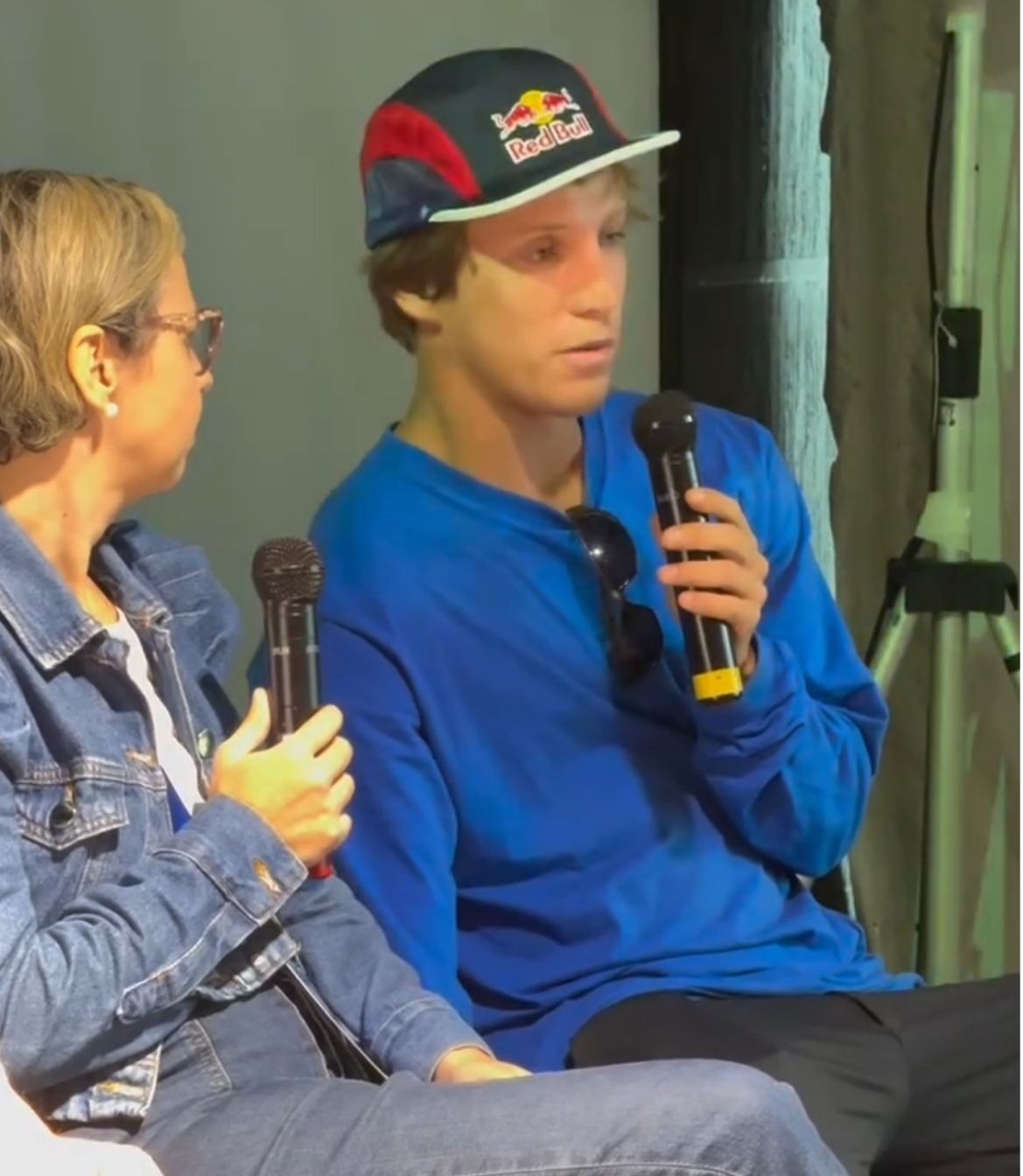Jovem com boné da Red Bull e camiseta azul fala ao microfone durante um painel, ao lado de uma mulher de jaqueta jeans que também segura um microfone