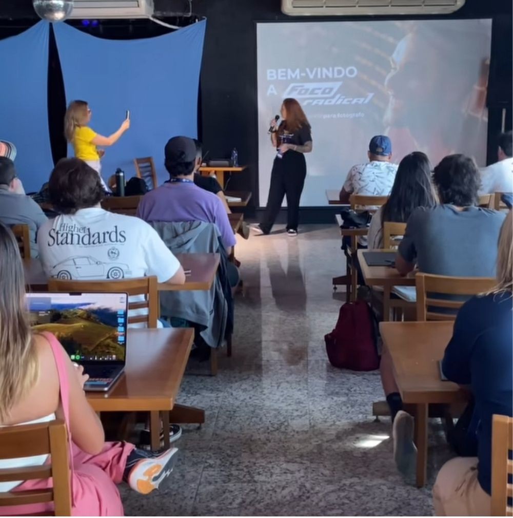 Palestrante com microfone diante de telão com "Bem-vindo a Foco Radical" e plateia sentada em mesas, alguns com notebooks, em um evento