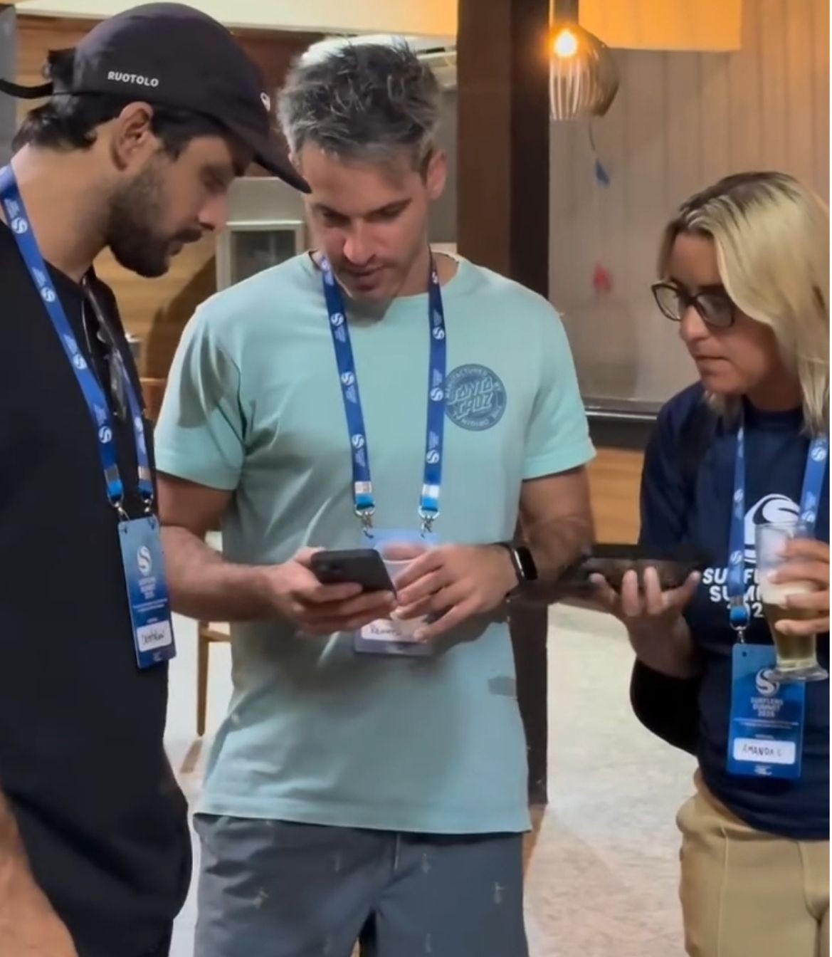 Dois homens e uma mulher com crachás do Surfers Summit olham juntos para o celular, em um ambiente interno; ela segura um copo de cerveja.